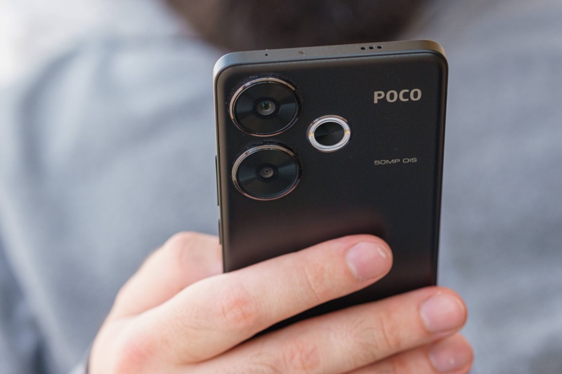 A hand holding the black Poco F7 5G smartphone with dual rear cameras and 50MP OIS label visible